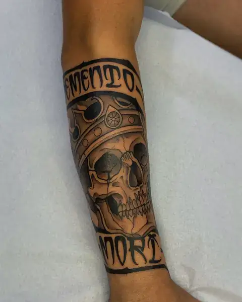 Tatuagem neotradicional de caveira com coroa e faixas “Memento mori” no antebraço